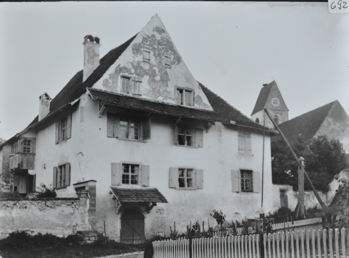 Mehrstöckiges gemauertes Wohnhaus, Fusswalmdach, kleines Dach oberhalb der Eingangstür, Steinmauer links und rechts neben dem Haus, Strasse vor dem Haus, hinten rechts weiteres Gebäude