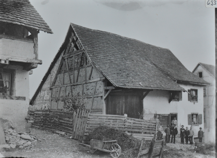 Mehrstöckiges Bauernhaus, grösstenteils gemauert, eine Wand aus Holz, Schleppdach, Menschengruppe posiert vor dem Hauseingang, im Vordergrund ein kleiner Weg sowie ein Holzaun mit Heu und Schubkarre 