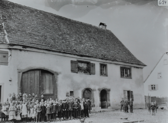 Zweistöckiges gemauertes Gebäude, Satteldach, vor einem grossen Tor posieren Kinder, weitere Kinder an der rechten Seite des Gebäudes