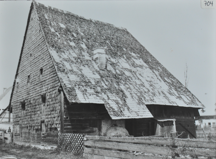 Mehrstöckiges Gebäude aus Holz, Fundament aus Stein, steiles Schleppdach mit Kamin, Feldweg mit Holzzaun vor dem Gebäude