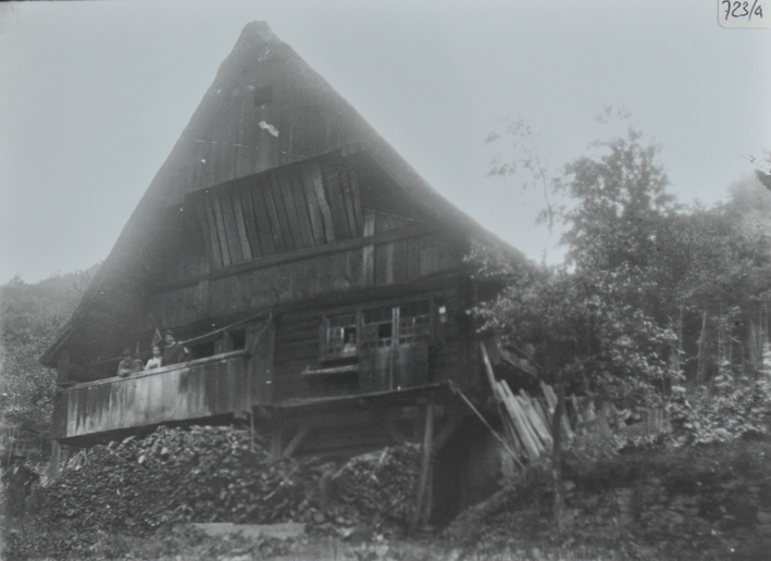Mehrstöckiges Bauernhaus aus Holz, Satteldach, auf dem Balkon posieren drei Personen, eine weitere Person schaut aus dem Fenster, vor dem Haus liegt gestapeltes Brennzolz, Sträucher rechts neben dem Haus