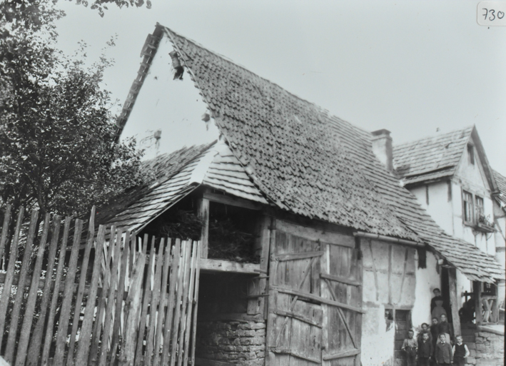 Zweistöckiges Bauernhaus mit Zwerchdach, vorderer Teil wird als Scheune genutzt, Scheunentor aus Holz, in der Scheune Brennzholz gelagert, vor dem Haus posiert eien Frau und mehrere Kinder