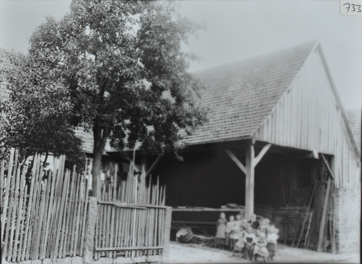 Bauernhaus mit Fundament aus Stein, oberer Teil aus Holz, Sattelach, welches auf der linken Seiten auf Holzpfeilen steht, mehrere Kinder posieren, links vom Haus ist ein Garten mit einem belaubten Baum, Gartenzaun aus Holz