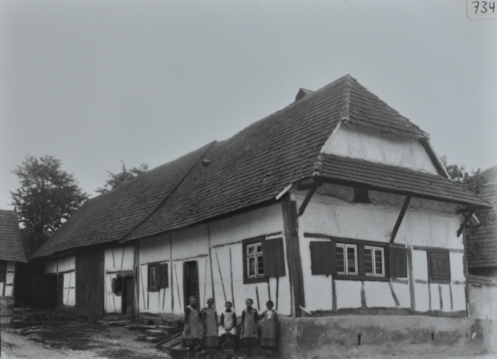 Bauernhaus gemauert, Fusswalmdach, Gelände leicht abfallend, vorderr Teil des Hauses steht auf einem gemauerten Fundament, beim Fundament posieren fünf Personen, Strasse links neben dem Haus