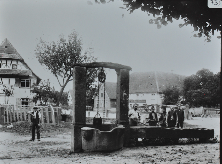 Ansicht eines Dorfplatzes, in der Mitte steht ein Ziehbrunnen, daneben posieren fünf Brunne, auf der Strasse posiert ein Mann mit einer Sense, im Hintergrund sind mehrere Gebäude 