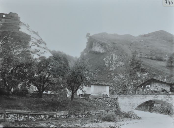Ansicht des Dorfes vom Fluss aus aufgenommen, Fluss fliesst durch das Dorf, eine Strasse und eine Steinbrücke verläuft quer durch das Dorf, im Hintergrund steht ein mehrstöckiges Bauernhaus aus Holz mit einem Satteldach, Hügel im Hintergrund