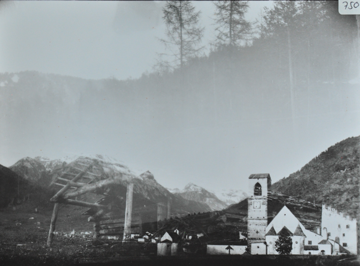 Doppelbelichtetes Bild mit einer Alphütte aus Holz sowie einem Dorfausschnitt mit mehreren Gebäuden und einer Kirche mit einem Kirchenturm, Bergkette im Hintergrund