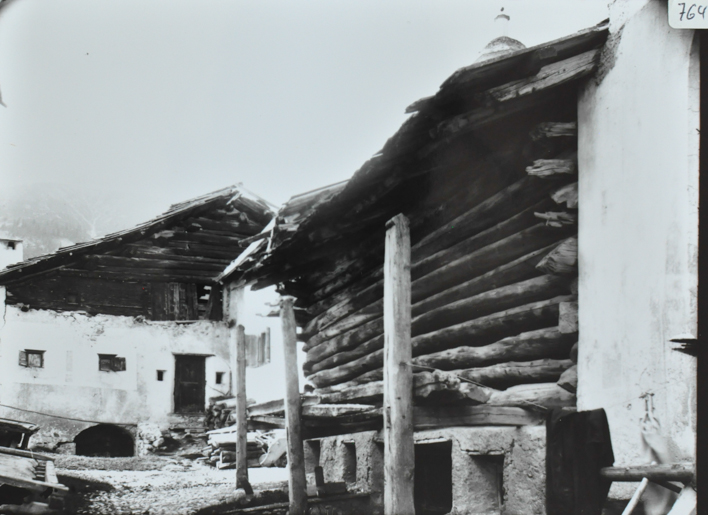 Wohnhaus aus Stein und Holz mit Satteldach, vor dem Haus gestapeltes Brennholz, im Vordergrund Speicher aus Holz mit Fundament aus Stein, Dach aus Holz, gepflasterte Strasse verbindet die Gebäude