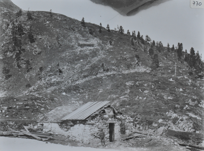 Aufnahme einer Alphütte aus Stein, Satteldach aus Stein und Holz, wobei eine Hälfte des Daches zerstört ist, Mann posiert vor der Hütte, Holzdielen liegen neben der Hütte, Hügel im Hintergrund