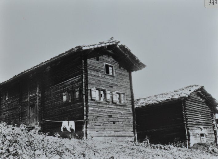 Ansicht von einem zweistöckigen Wohnhaus aus Holz, Satteldach Wäscheleine vor dem Haus aufgehängt, neben dem Wohnhaus befindet sich ein weiteres ähnliches Gebäude aus Holz und mit Satteldach, allerdings ohne Fenster