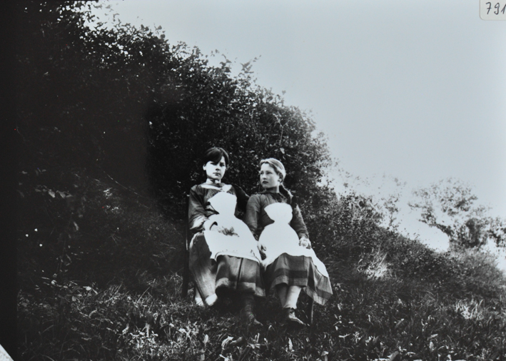 Zwei Mädchen posieren auf den auf der Wiese sitzend auf einem Stuhl, Gebüsch im Hintergrund