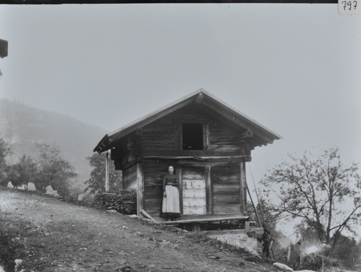 Alphütte aus Holz, mit Fundament aus Stein, zwischen Fundament und Haus sind Pflöcke, Haus am Hang erbaut, Frau posiert vor der Eingangstür, links an der Wand liegt Brennholz