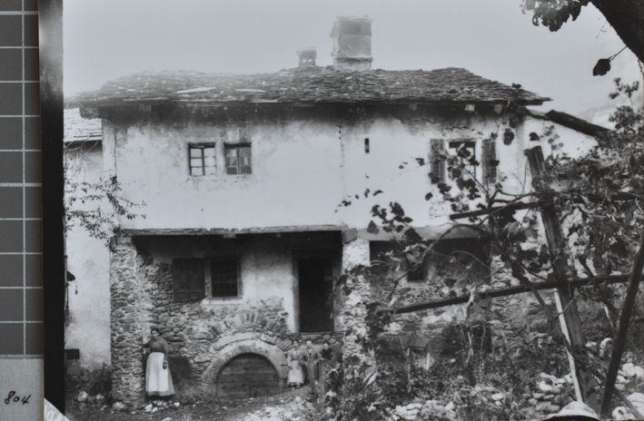 Zweistöckiges Wohnhaus aus Stein, obere Etage verputzt, Satteldach mit Kamin, vor dem Haus posiert eine stehenden Frau sowie drei sitzende Personen, Garten vor dem Haus