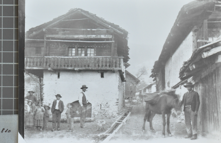 Bauernhaus mit Fundament aus Stein, oberer Teil aus Holz, Satteldach, vor dem Haus posieren Ewachsenen mit Kindern, auf der Strasse, welche durch das Dorf führt, posiert ein Mann mit einem Esel, weitere Gebäude an Strasse angebaut