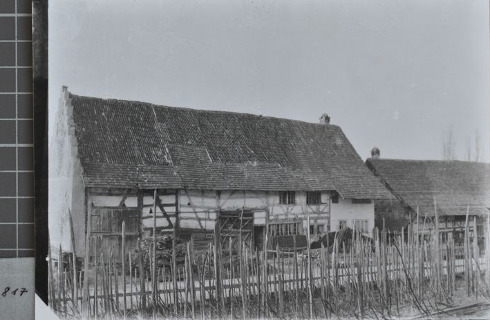 Gemauertes Bauernhaus mit Schleppdah, Scheunentor aus Holz, vor dem Haus liegt gestapeltes Brennholz, vor dem Haus führt eine Strasse vorbei, rechts ist ein weiteres Bauernhaus mit Schleppdach