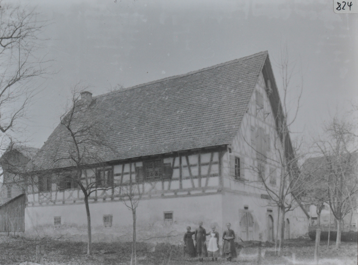 Mehrstöckiges gemauertes Wohnhaus, Satteldach mi Kamin, vier Personen posieren auf der Wiese vor dem Haus, mehrere kahle Bäume vor dem Haus, im Hintergrund weitere Gebäude ersichtlich