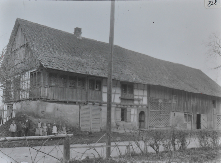 Zweistöckiges Bauernhaus, teils gemauert teils aus Holz, langes Satteldach, mehrere Kinder posieren vor dem Haus, dahinter Misthaufen, vor dem Haus verläuft Strasse