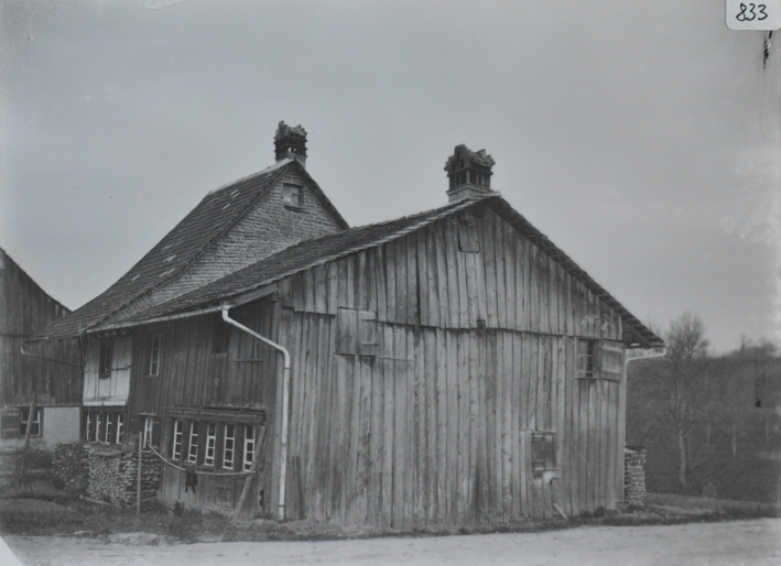 Zweistöckiges Bauernhaus mit angebauter Scheune, Haus und Scheune mit Satteldach und Kamin, an den Gebäuden liegt gestapeltes Brennholz, Strasse vor der Scheune, im Hintergrund weiteres Gebäude 