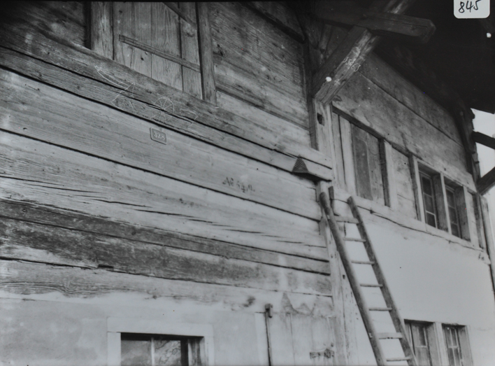 Ansicht einer Hausfassade aus Holz, Holzleiter an die Fassade angelegt, Teil des Daches ersichtlich, geritzte Formen in der Fassade