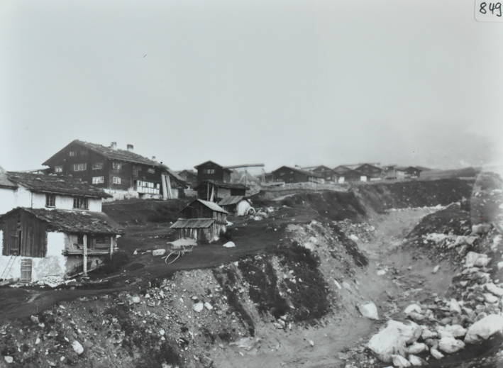 Ansicht eines Dorfausschnittes mit mehreren Gebäuden, Fundament gemauert, Gelände nach hinten steigend, Grosse Grube vor den Gebäuden