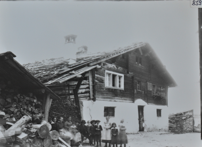 Zweistöckiges Bauernhaus mit Fundament aus Holz, oberer Teil aus Holz, Schleppdach, rechts neben dem Gebäude liegt gestapeltes Brennholz, davor poiseren mehrere Kinder