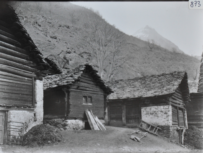 Ansicht eines Dorfteils mit mehreren Gebäuden aus Holz und Fundament aus Stein, Gebäude einstöckig, Satteldächer, Weg verbindet die Häuser, Berg im Hintergrund