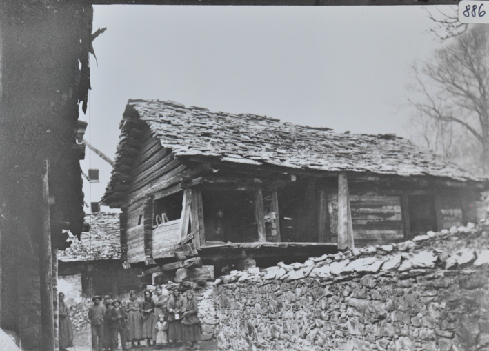 Einstöckiges Wohnhaus aus Holz stehend auf Pfeilern aus Stein und Holz, Satteldach aus Stein, neben dem Haus verläuft Steinmauer, daneben posiert eine Menschengruppe, weitere Gebäude im Hintergrund