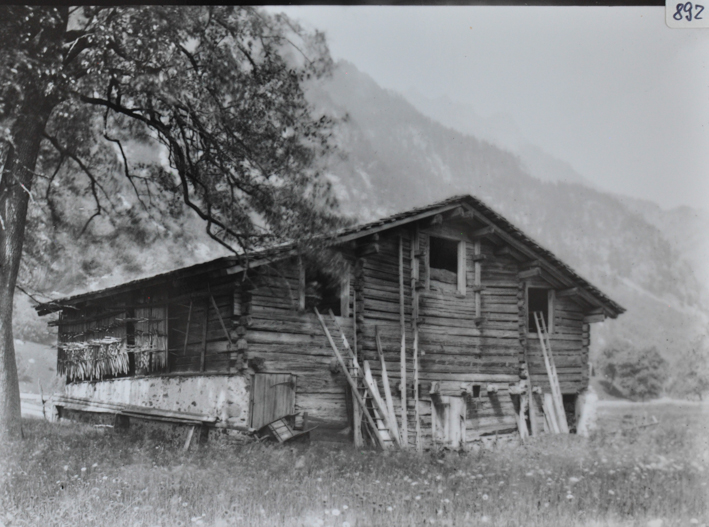 Scheune aus Holz mit Satteldach, Fundament gemauert, vor der Scheune liegen Leiter und weitere Gegenstände, Scheuen steht auf einer Wiese, Hügel im Hintergrund