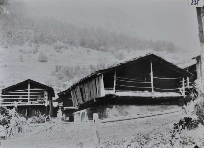 Ansicht eines zweistöckigen Wohnhauses aus Holz sowie eine Speichers aus Holz, Speicher auf Fundament und Stützen aus Stein erbaut, Weg führt durch das Dorf, Hügel im Hintergrund