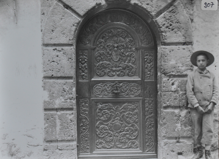 Ansicht eines Tors aus Metall, Tor verziert, Steinelemente um das Tor, Kind mit Hut possiert an der Wand