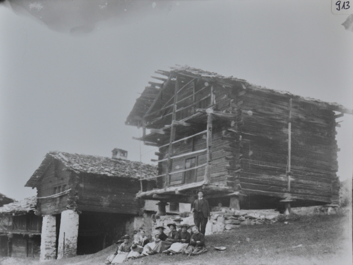 Ansicht von zwei Wohnhäusern aus Holz, Satteldächer, Häuser stehen auf Pfeilern resp. auf Fundament aus Stein aufgrund des abfallenden Geländes, mehrer Personen posieren vor dem ersten Haus