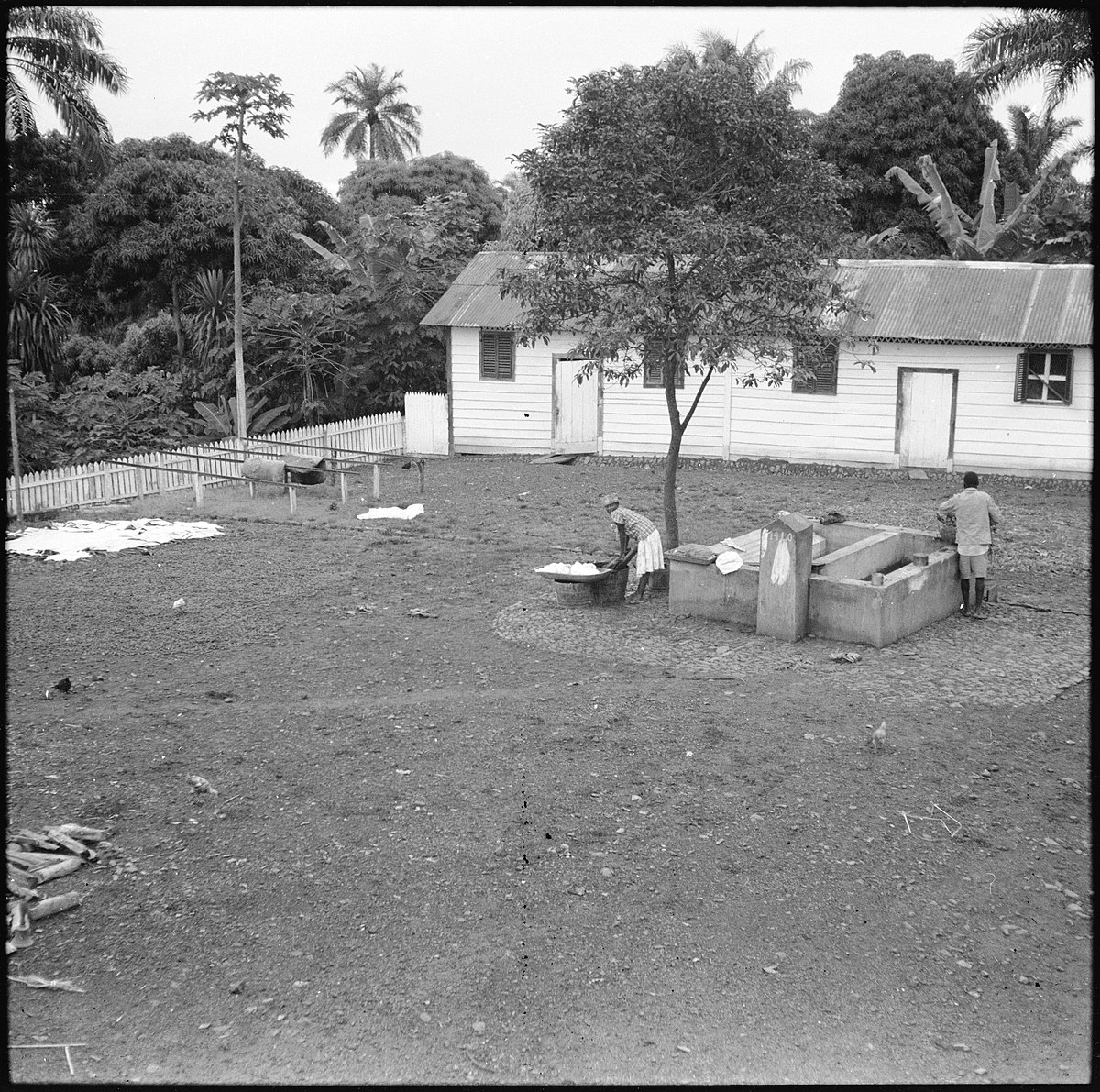 Portugal, San Thomé (São Tomé und Príncipe): Landschaft; Baracke im Hintergrund; Frau und Mann auf offenem Platz am waschen