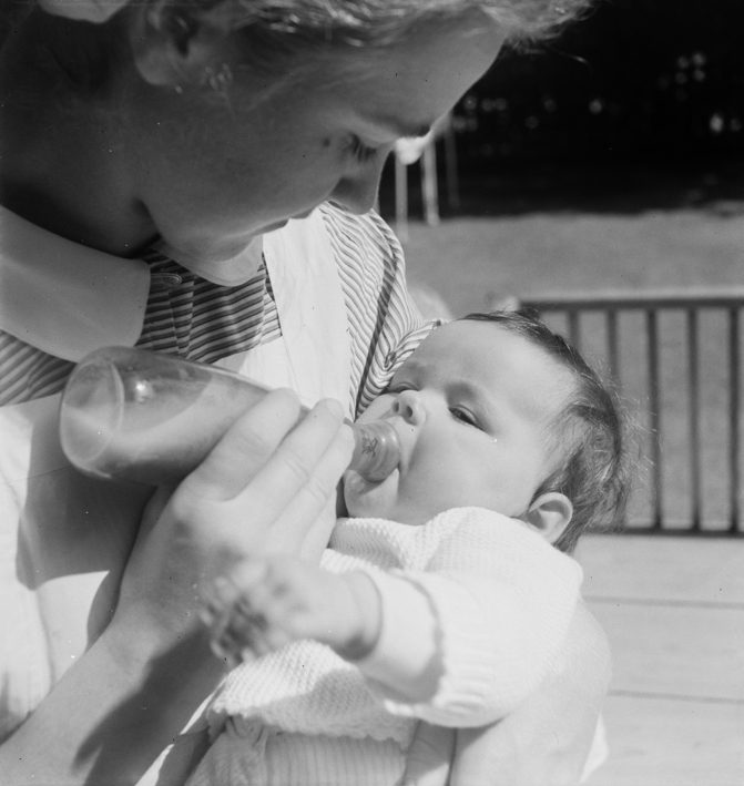 Krankenschwester in Tracht gibt einem Baby den Schoppen auf einer sonnigen Terrasse