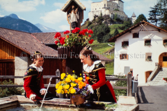 Zwei Frauen in Trachten mit Blumen bei Brunnen, Schloss Tarasp im Hintergrund
