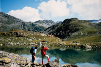 Zwei Personen beim Wandern neben Bergsee, Kuhherde gegenüber
