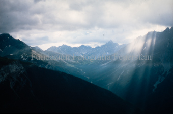 Blick von Bergen ins Tal und auf Alpen, bedeckter Himmel mit Strahlenbüschel