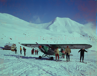 Propellerflugzeug (vermutlich Piper PA-20), Schneelandschaft, Skifahrer