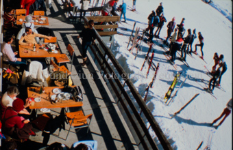 Personen auf Restaurantterrasse neben Skipiste mit Skifahrer