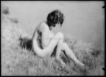 Frauenakt sitzend im Gras, an einem See, Arme vor dem Oberkörper verschränkt, Blick nach unten