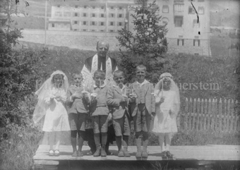 Gruppenaufnahme, Erstkommunion, zwei Mädchen, drei Knaben und ein Geistlicher, im Hintergrund grosses Gebäude