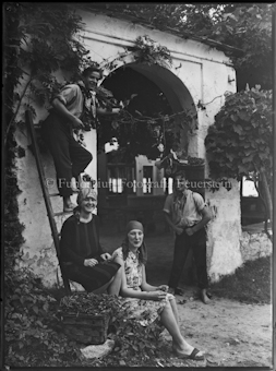 Zwei Frauen und Männer stehend und sitzend vor und auf Leiter vor Torbogen