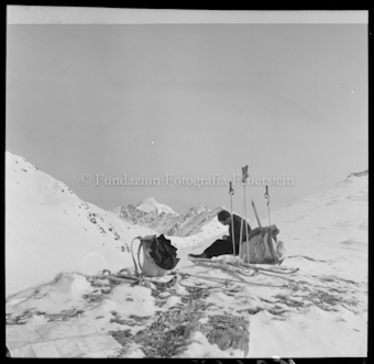 Person mit Ski, Stöcken und Rucksäcken sitzend
