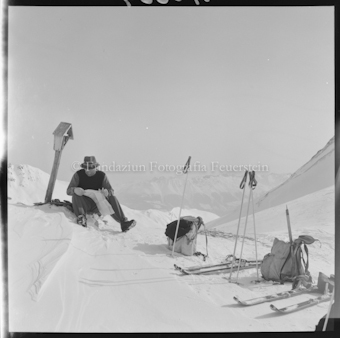 Person mit Ski, Stöcken und Rucksäcken sitzend