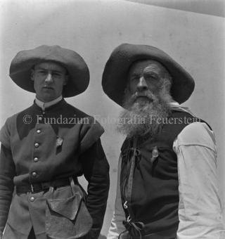 Jüngerer Mann neben älterem Mann mit Bart, beide mit Hut und angestecktem Emblem in historischen Kleidern