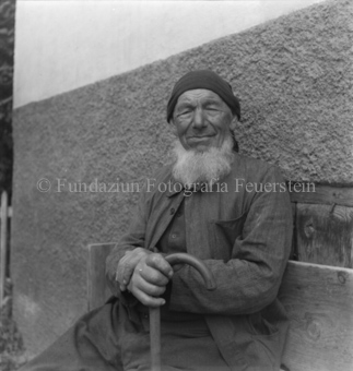 Älterer Mann mit Bart und Mütze, sitzend mit Gehstock in Hand
