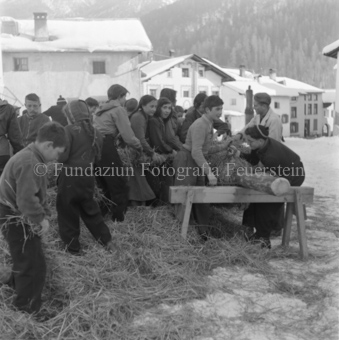 Kinder in Strohhaufen auf Dorfplatz, Stroh um Stamm wickelnd