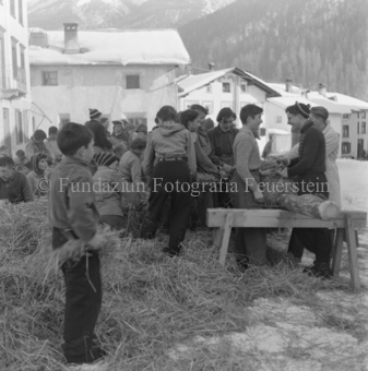 Kinder in Strohhaufen auf Dorfplatz, Stroh um Stamm wickelnd