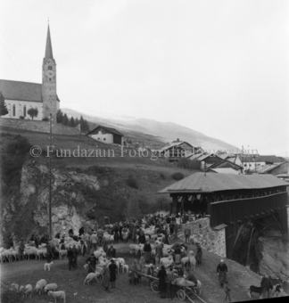 Schafherde und Menschen bei Brücke, hinten Dorf und Kirche
