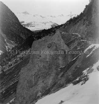 Grosser Fels an Berghang, oben abgezäunter Weg, teilweise Schnee