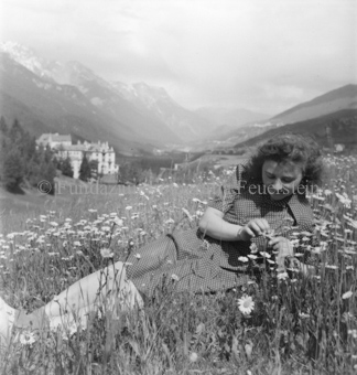 Frau in Blumenwiese liegend, Tal und Gebäude im Hintergrund